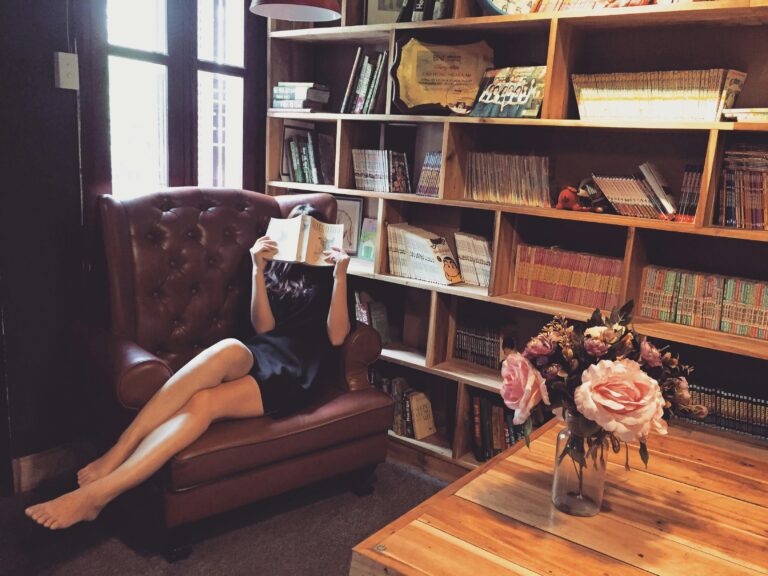 Woman relaxing with a book in a cozy library armchair, surrounded by wooden bookshelves and flowers.